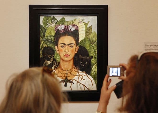 A picture of Frida Kahlo's "Self-Portrait with Thorn Necklace and Hummingbird" taken as it hangs in the museum, two museum goers standing in front of it.